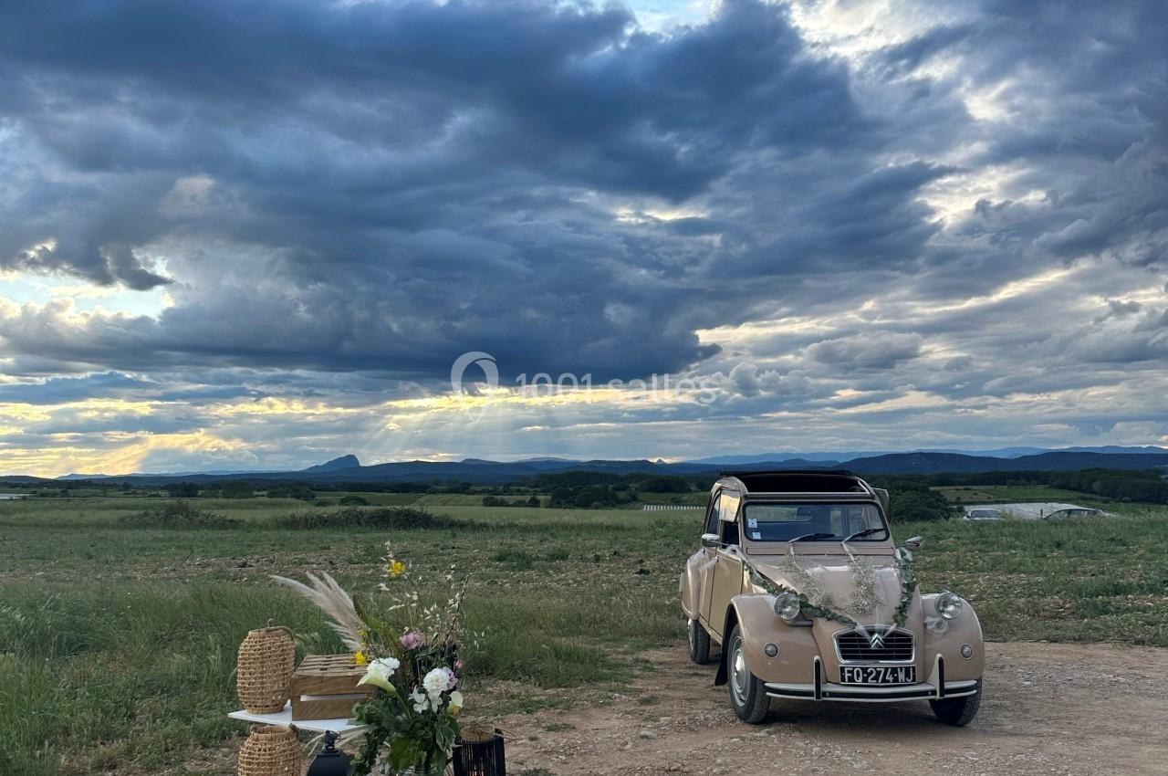 Une voiture ancienne beige garée dans un paysage rural sous un ciel nuageux, avec une petite décoration champêtre au premier…