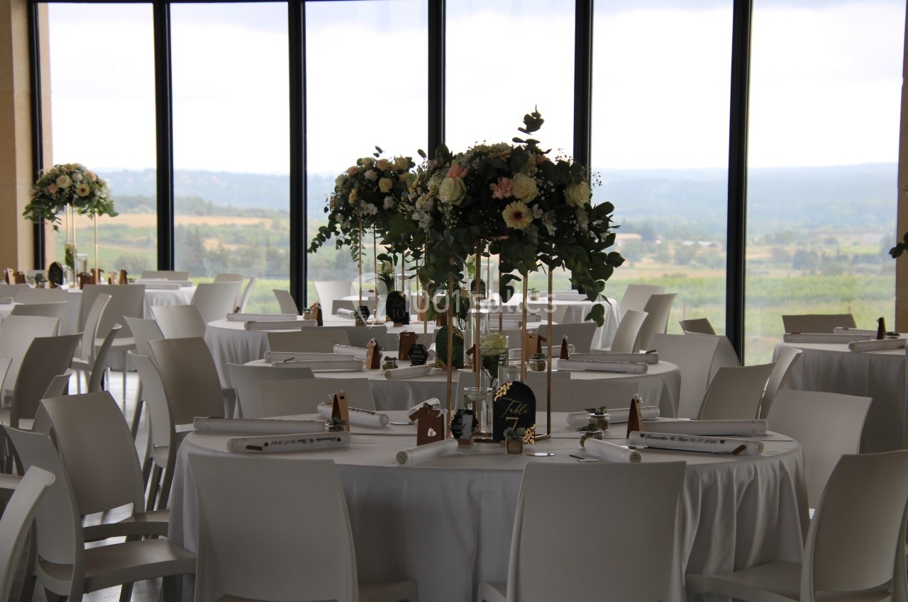 Salle de réception lumineuse avec tables rondes décorées de nappes blanches, fleurs et chandeliers, vue sur la campagne.