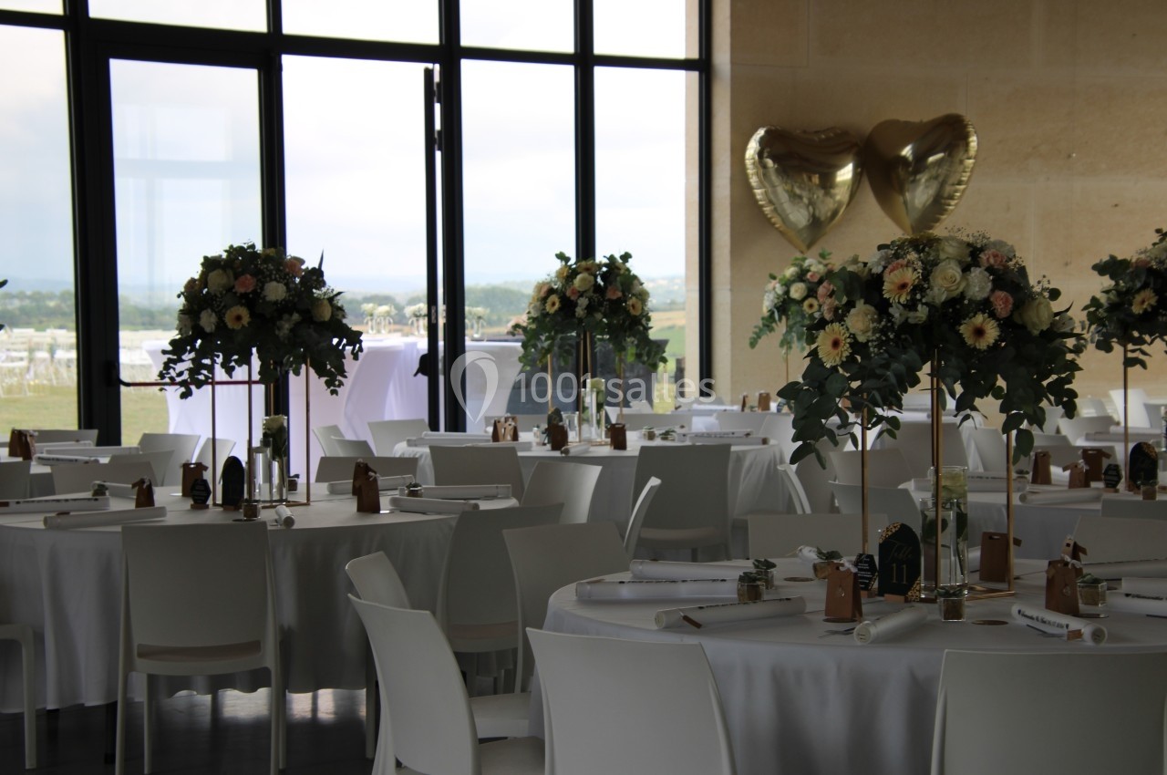 Salle de réception décorée pour un mariage, avec tables rondes, bouquets de fleurs et ballons dorés en forme de cœur.