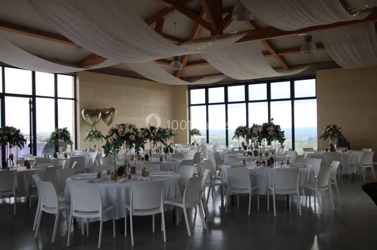 Salle de réception décorée avec des tables rondes, nappes blanches, fleurs et ballons dorés en forme de cœur.