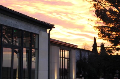 Façade vitrée d'un bâtiment moderne au coucher du soleil, entourée d'arbres et d'un ciel aux teintes orangées.