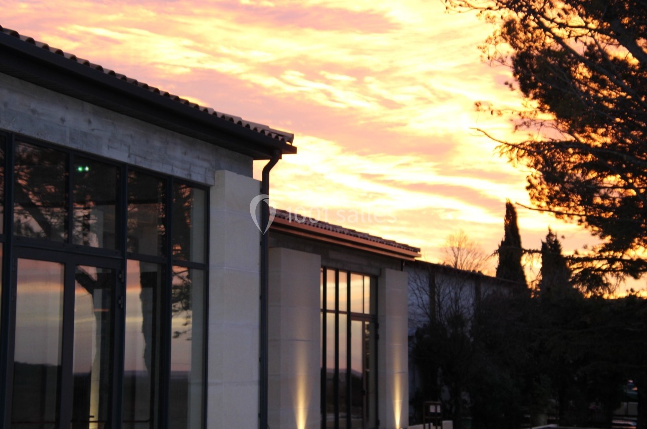 Façade vitrée d'un bâtiment moderne au coucher du soleil, entourée d'arbres et d'un ciel aux teintes orangées.