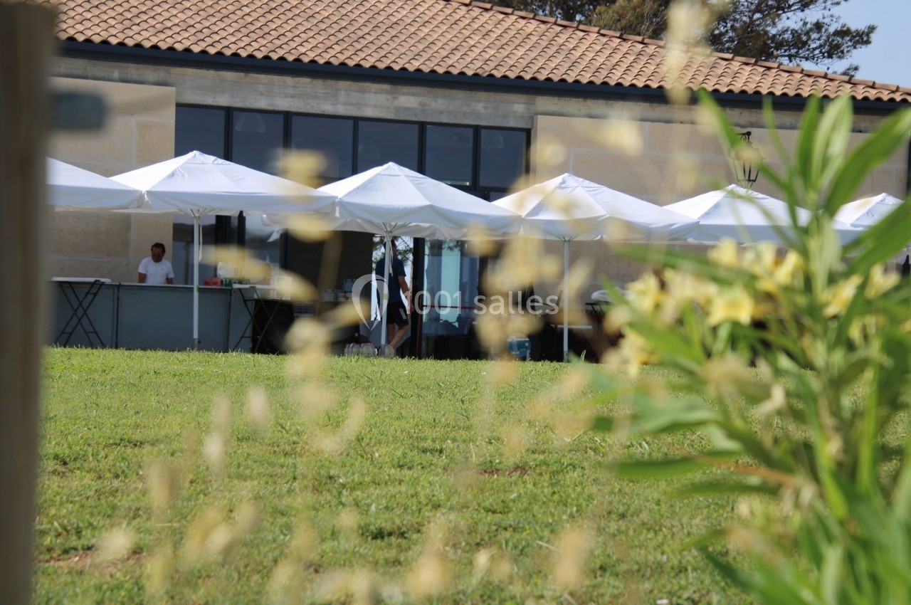 Terrasse avec parasols blancs devant un bâtiment vitré, vue à travers des herbes floues au premier plan.