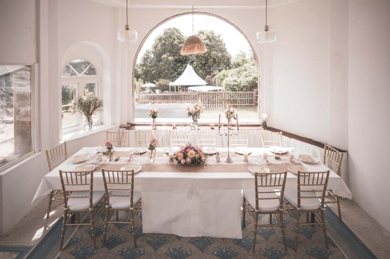 Table élégamment dressée avec des chaises dorées, fleurs au centre et vue sur un jardin à travers une grande fenêtre.