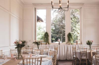 Salle lumineuse décorée pour un événement, avec des tables dressées, des chaises dorées et des fleurs en centre de table.
