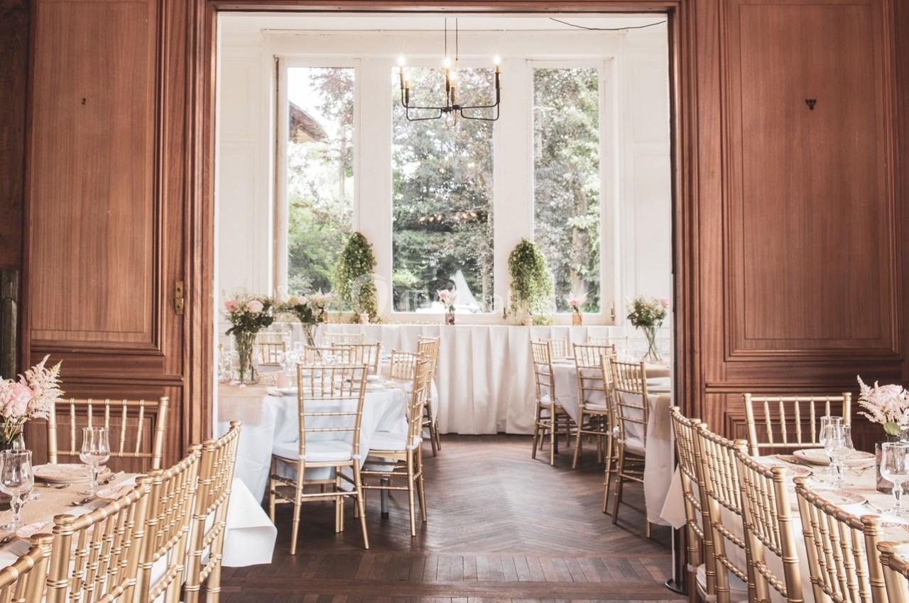 Salle de réception élégante avec tables dressées, chaises dorées et vue sur un jardin à travers de grandes fenêtres.