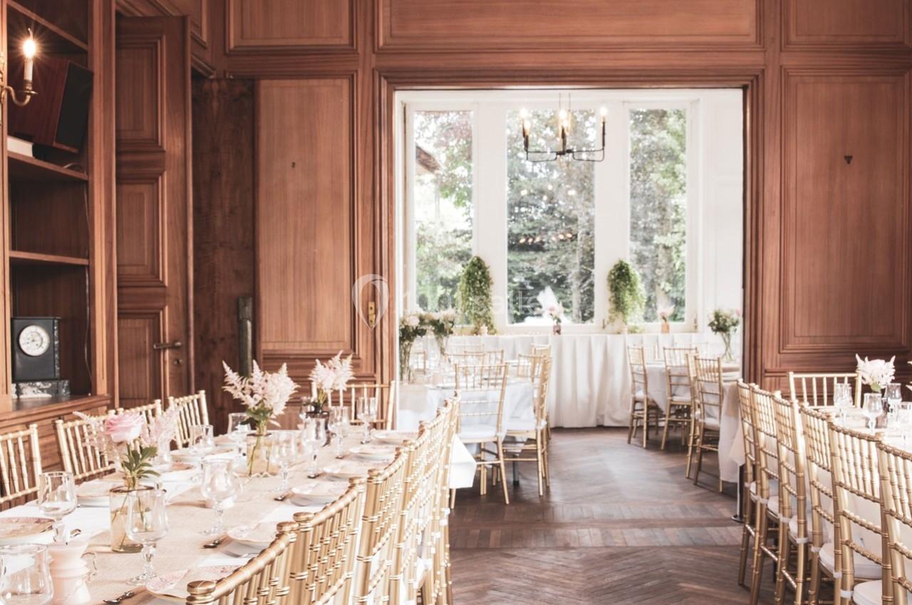 Salle de réception élégante avec tables dressées, chaises dorées et grandes fenêtres donnant sur un jardin verdoyant.