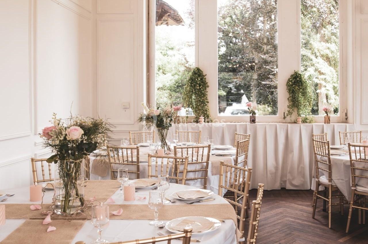 Salle de réception lumineuse avec tables décorées de nappes blanches, fleurs roses et chaises dorées, vue sur jardin.