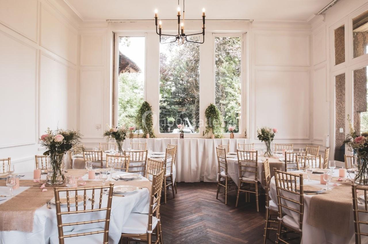 Salle lumineuse décorée pour un événement, avec des tables dressées, des chaises dorées et des fleurs en centre de table.