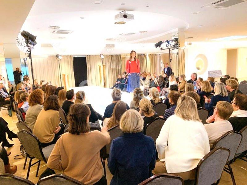 Défilé de mode dans une salle lumineuse avec une femme sur le podium et un public assis observant.