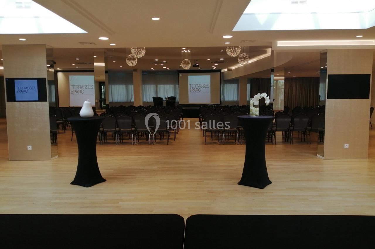 Salle de conférence avec rangées de chaises, deux tables hautes décorées et écrans affichant ’Terrasses du Parc’.