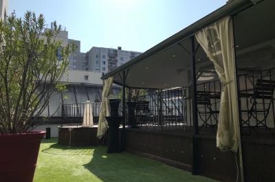 Terrasse extérieure avec pelouse artificielle, pergola couverte et immeubles en arrière-plan par temps ensoleillé.