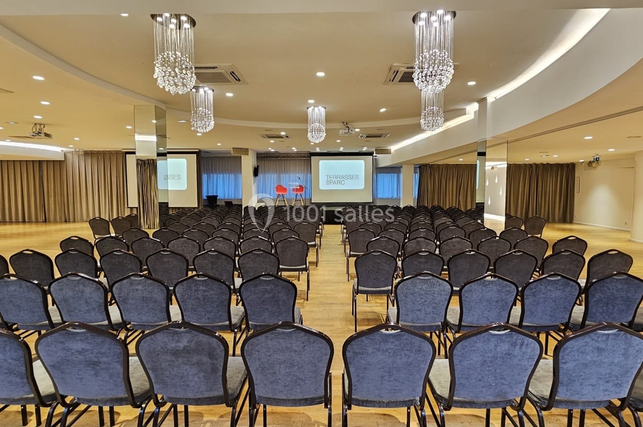 Salle de conférence avec rangées de chaises noires alignées face à une scène équipée de deux écrans et deux chaises rouges.