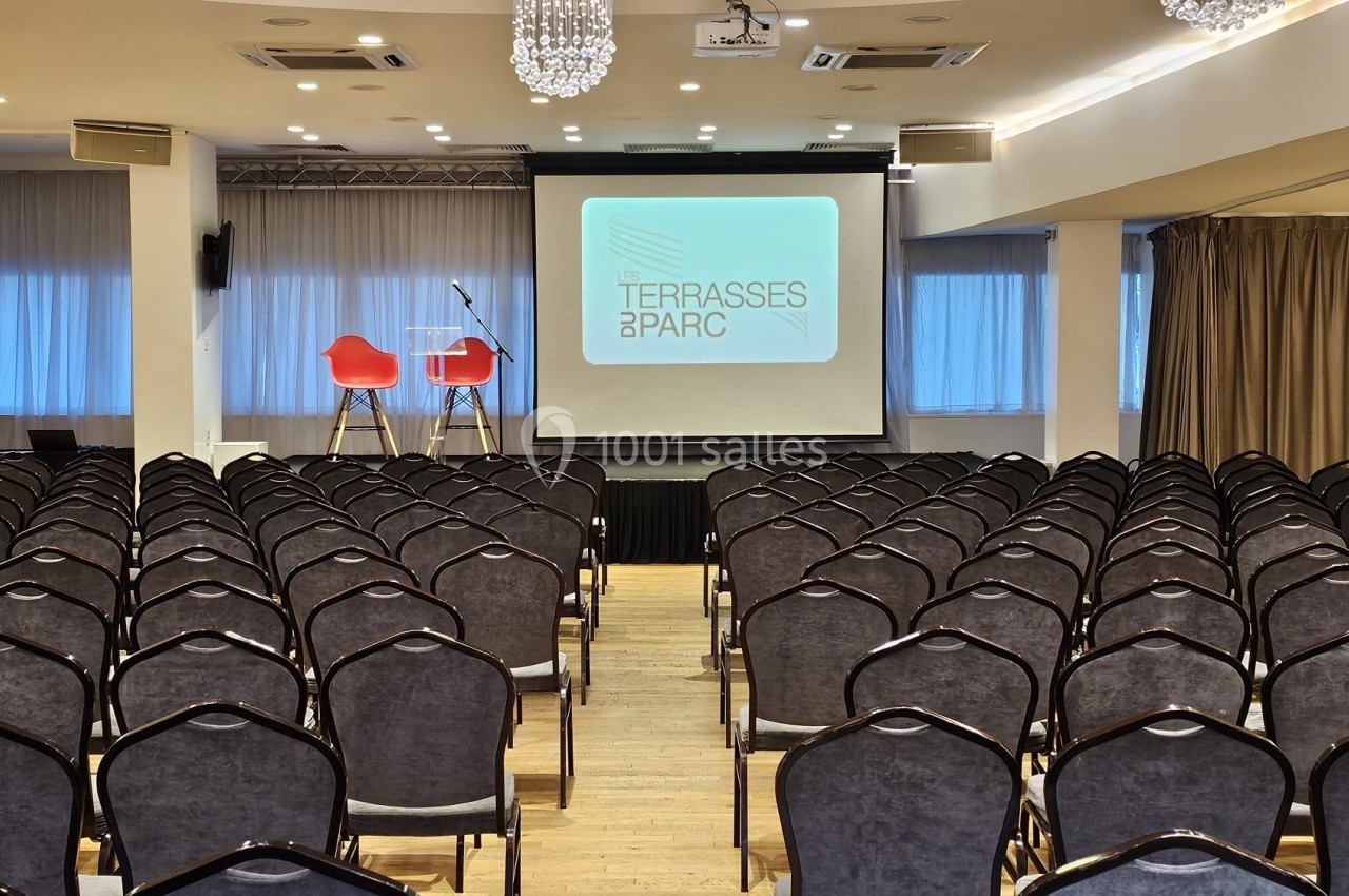 Salle de conférence avec rangées de chaises alignées, une scène équipée de deux chaises et un écran de projection.