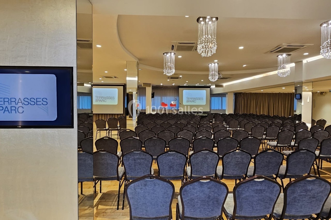 Salle de conférence avec rangées de chaises alignées, écrans affichant ’Les Terrasses du Parc’ et lustres suspendus.