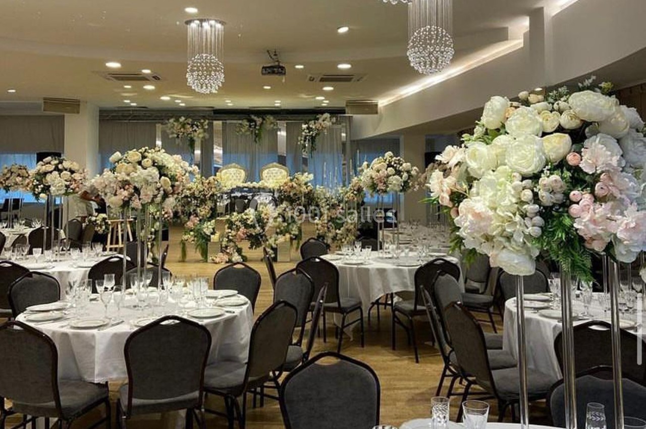 Salle de réception élégante décorée avec des tables rondes, nappes blanches et arrangements floraux raffinés.