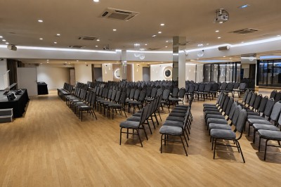 Salle de conférence avec des rangées de chaises noires alignées face à une scène et un écran de projection.