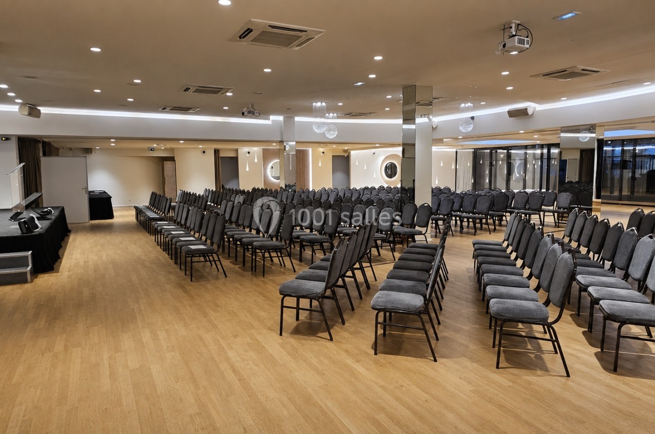 Salle de conférence avec rangées de chaises grises alignées, une scène à gauche et un éclairage moderne au plafond.