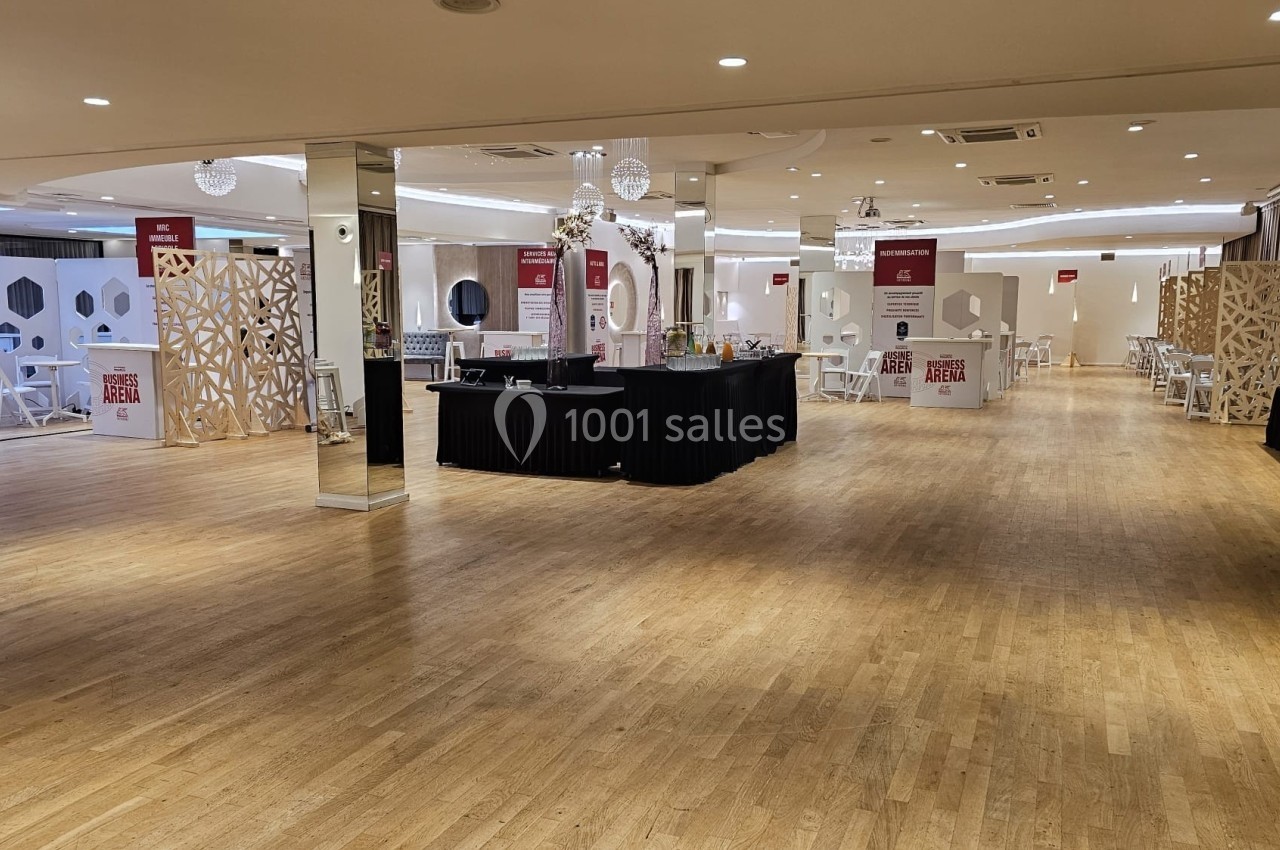 Salle spacieuse avec parquet clair, stands d'exposition, tables et chaises, éclairée par des plafonniers modernes.