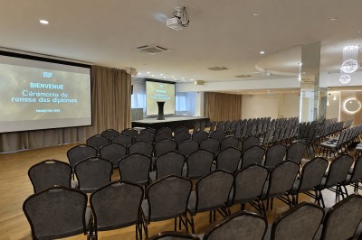 Salle de conférence avec des rangées de chaises noires alignées face à une scène et un écran de projection.