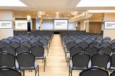 Salle de conférence avec des rangées de chaises noires alignées face à une scène et un écran de projection.