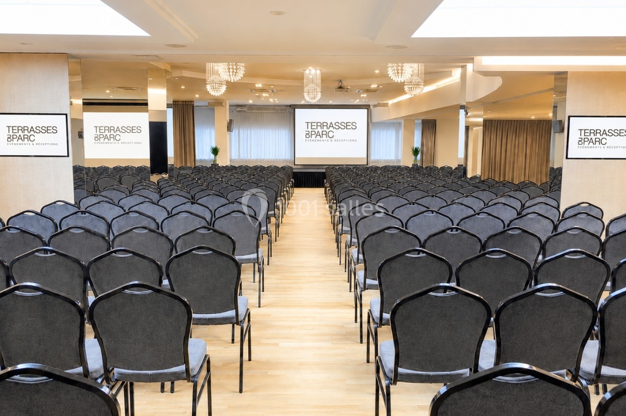 Salle de conférence avec rangées de chaises noires alignées face à deux écrans de projection et un espace lumineux.
