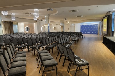 Salle de conférence avec des rangées de chaises noires alignées face à une scène et un écran de projection.