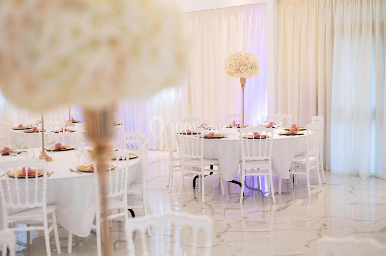 Salle de réception élégante avec tables rondes dressées, nappes blanches et décorations florales raffinées.