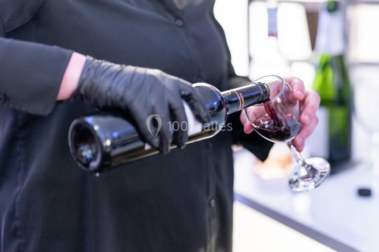 Une personne verse du vin rouge dans un verre à pied, portant des gants noirs, avec des bouteilles en arrière-plan.