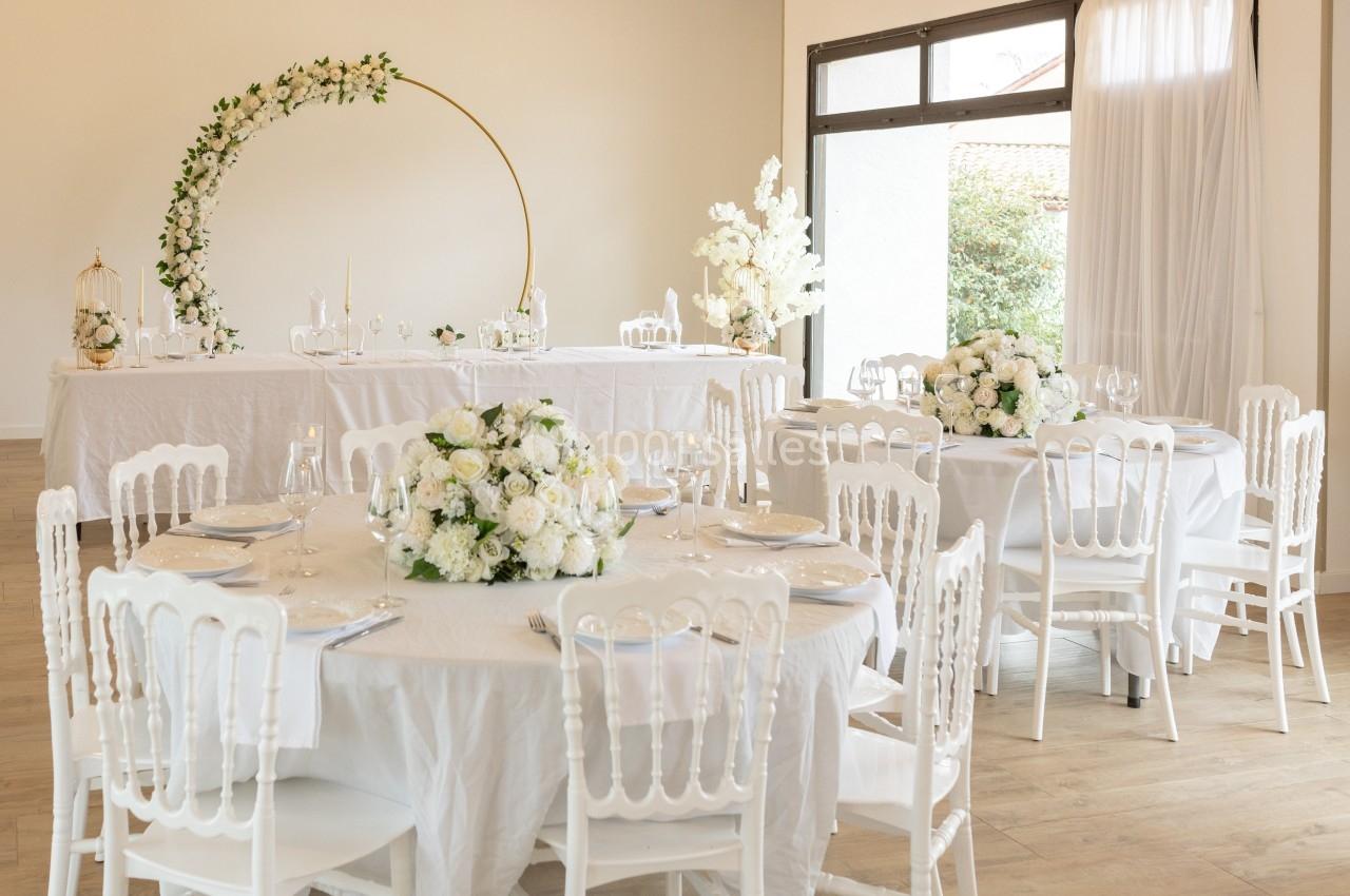 Salle de réception lumineuse décorée de tables rondes avec nappes blanches, chaises élégantes et arrangements floraux.