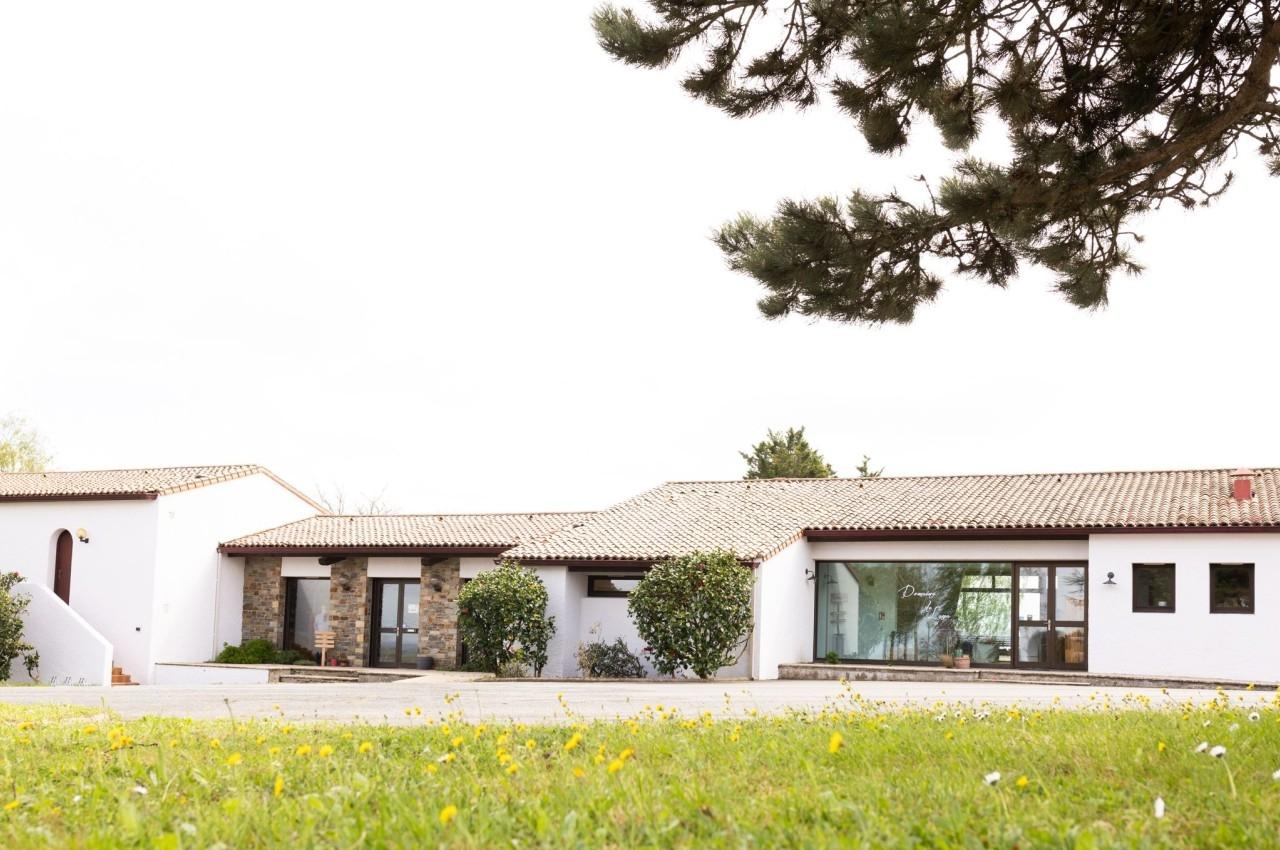 Bâtiment de plain-pied avec murs blancs, toit en tuiles et grandes baies vitrées, entouré de verdure et d'arbres.