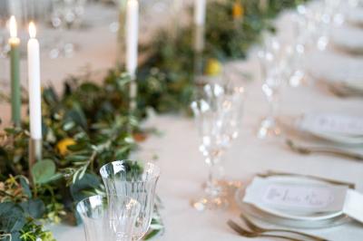 Table élégamment dressée avec nappes blanches, verres, couverts, bouquets de fleurs et petits pots décoratifs.
