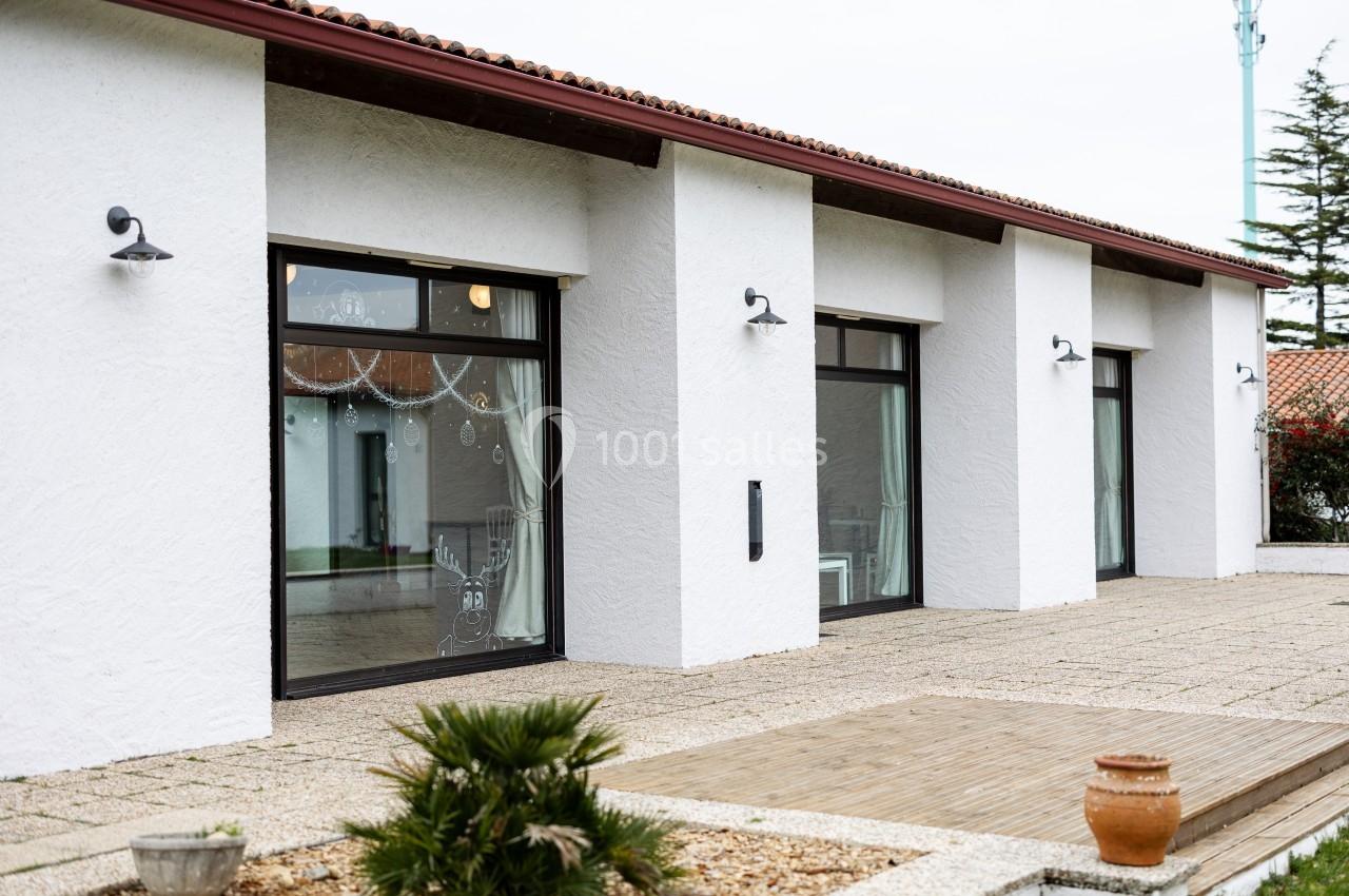 Façade d'une maison blanche avec grandes baies vitrées, éclairages muraux et terrasse en dalles et bois.