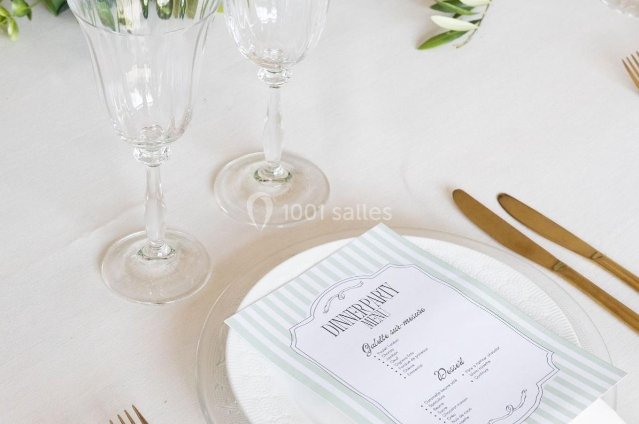Table dressée avec des couverts dorés, deux verres à pied, une assiette avec un menu et une décoration végétale.