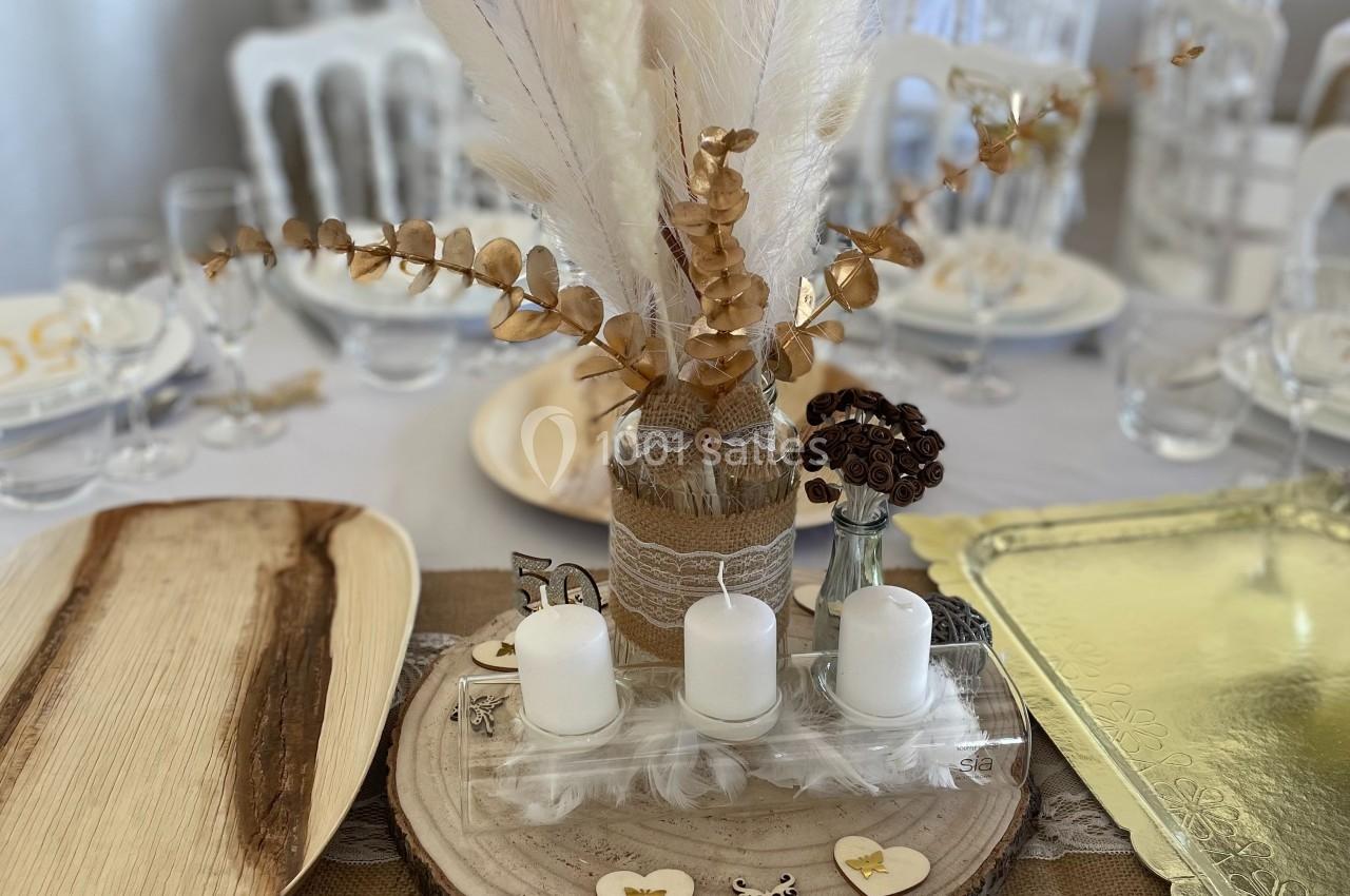 Centre de table avec bougies blanches, fleurs séchées et éléments décoratifs sur un rondin de bois, entouré de vaisselle.