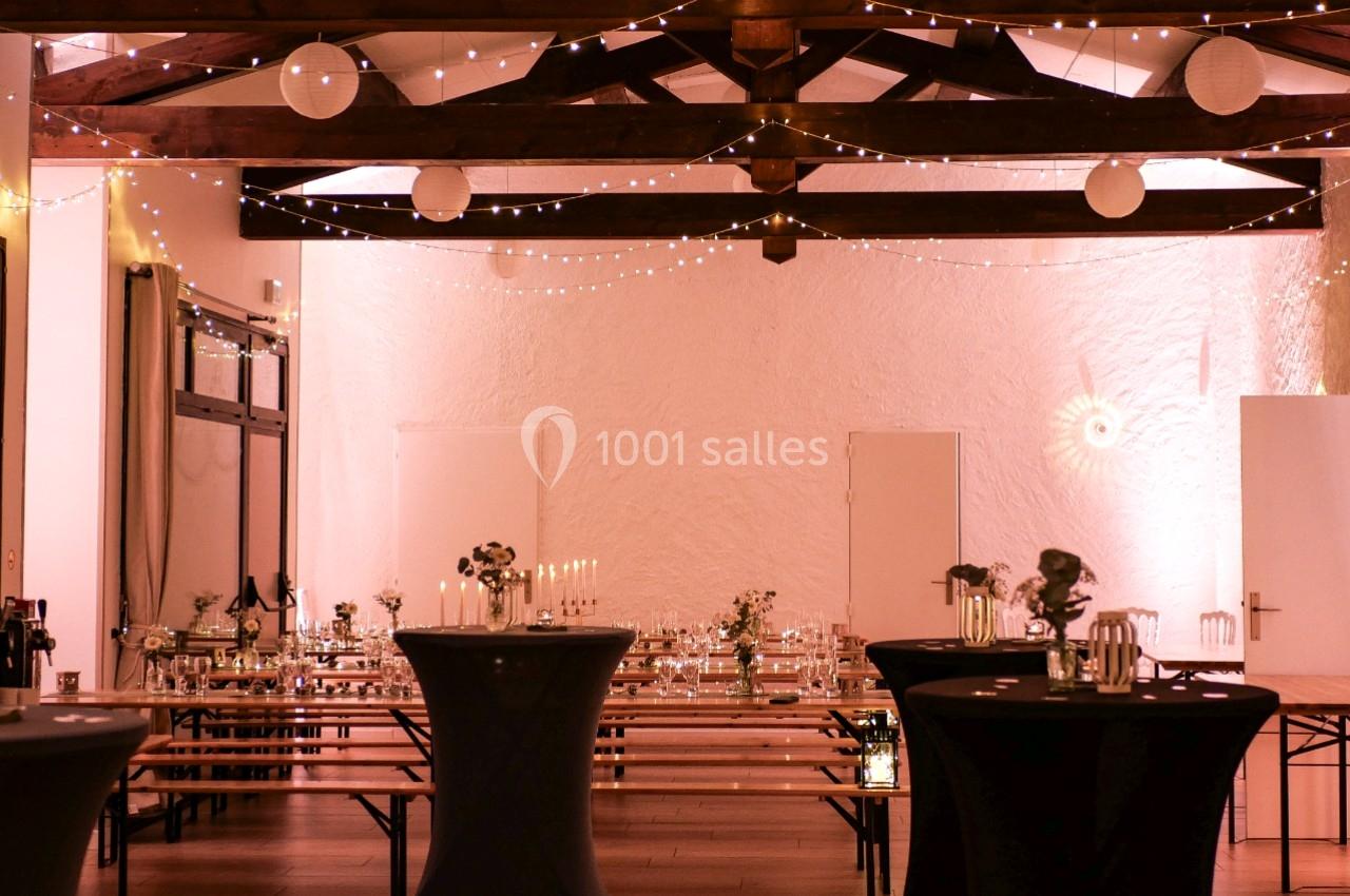 Salle décorée pour un événement, avec guirlandes lumineuses, tables hautes et bancs en bois sous une charpente apparente.