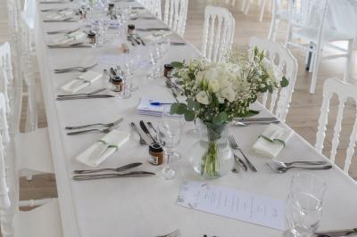 Table élégamment dressée avec nappes blanches, verres, couverts, bouquets de fleurs et petits pots décoratifs.