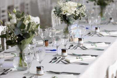 Table élégamment dressée avec nappes blanches, verres, couverts, bouquets de fleurs et petits pots décoratifs.