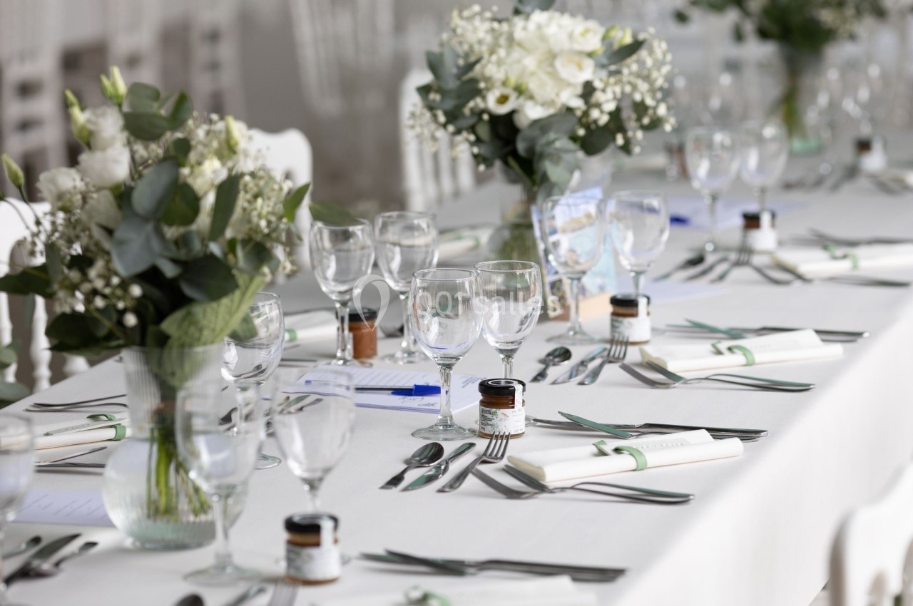 Table élégamment dressée avec nappes blanches, verres, couverts, bouquets de fleurs et petits pots décoratifs.