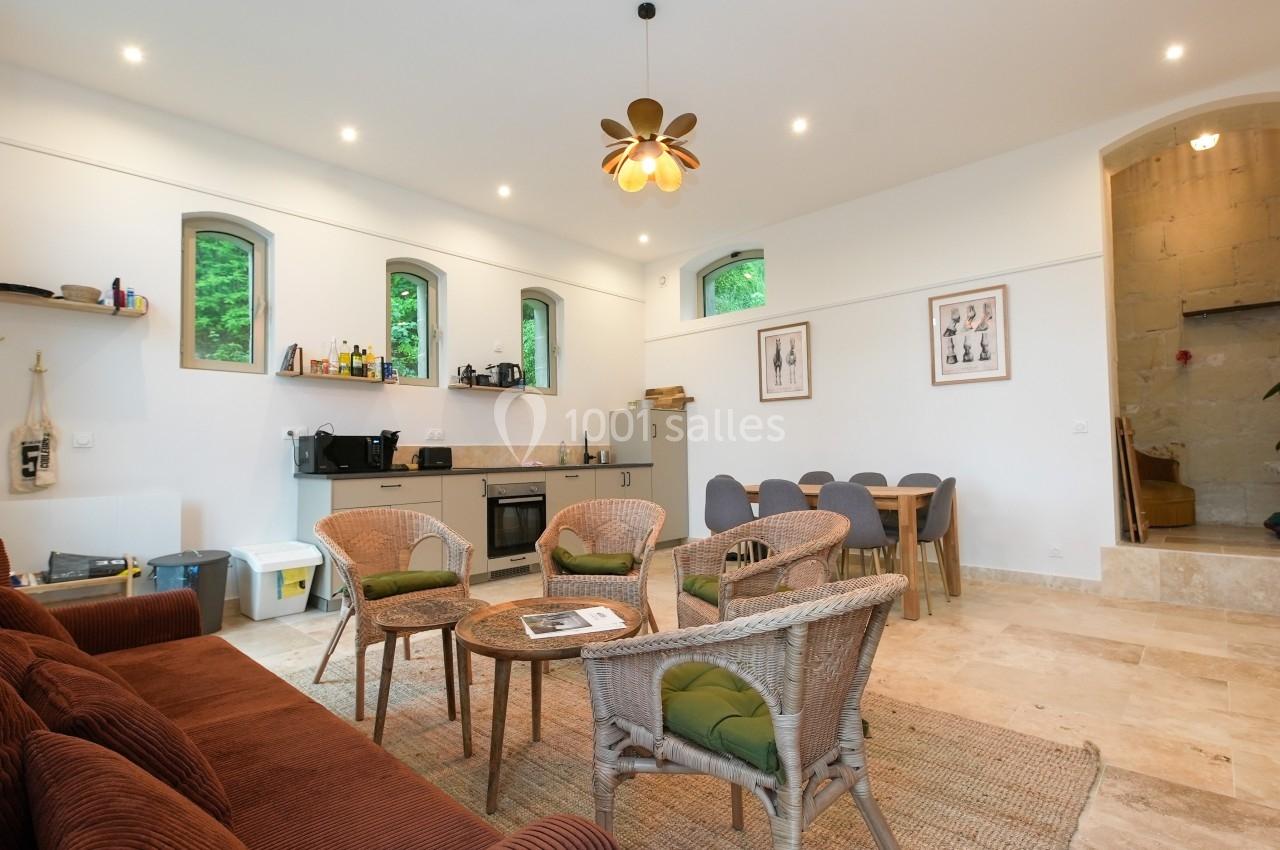 Salon lumineux avec canapé, fauteuils en osier, table basse, cuisine ouverte équipée et table à manger.