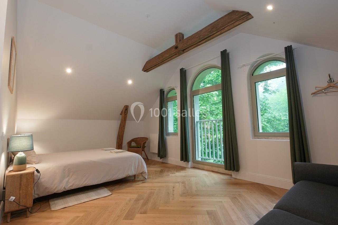 Chambre lumineuse avec lit double, parquet en bois, poutres apparentes, grandes fenêtres et rideaux verts.