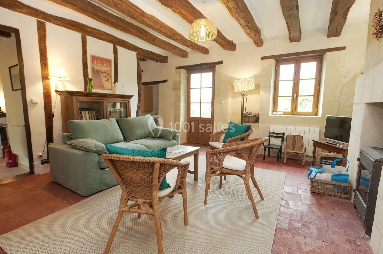 Salon lumineux avec poutres apparentes, canapé vert, fauteuils en osier, tapis clair et fenêtres donnant sur l'extérieur.