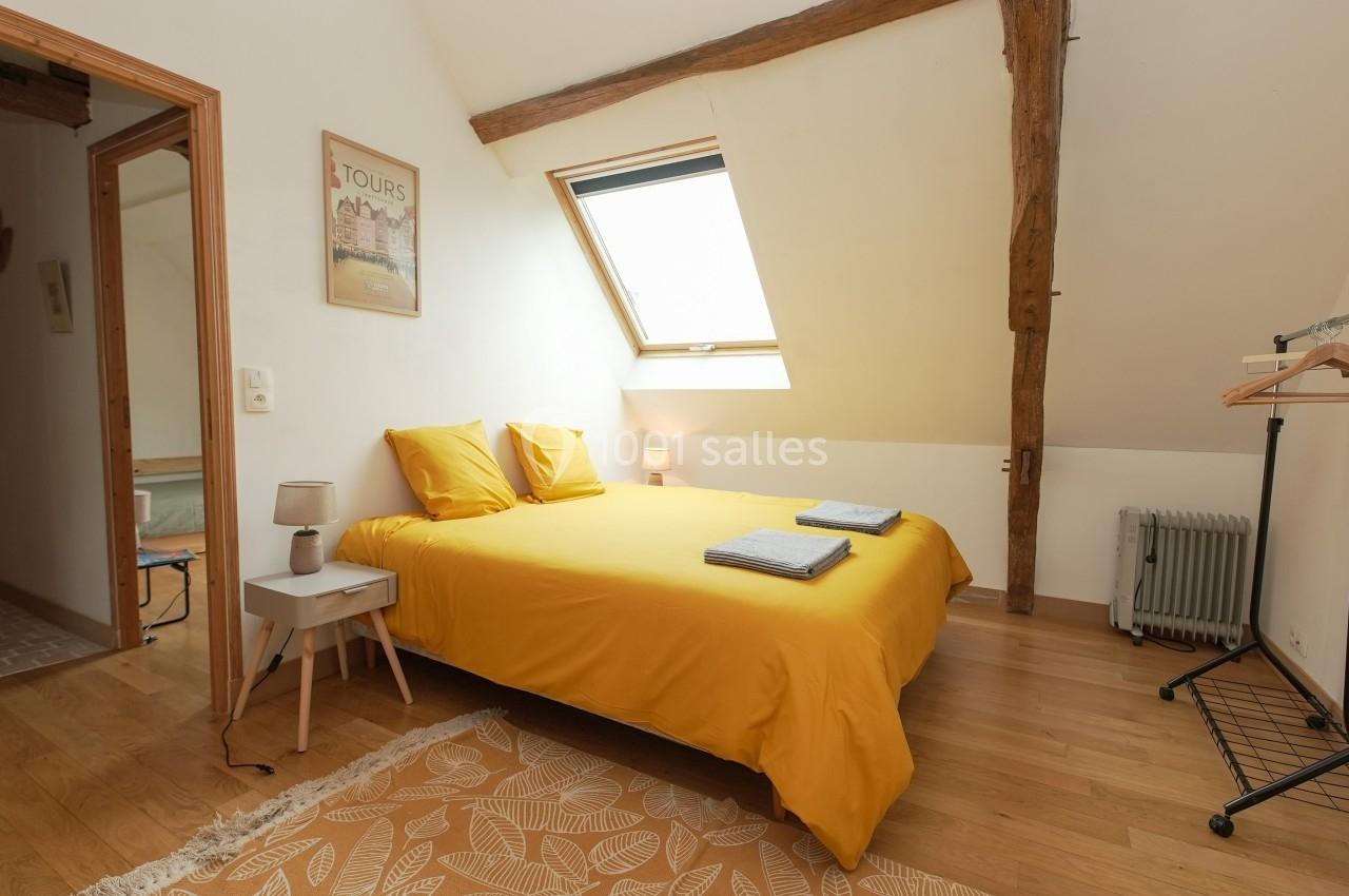Chambre mansardée lumineuse avec lit double aux draps jaunes, parquet en bois et fenêtre de toit.