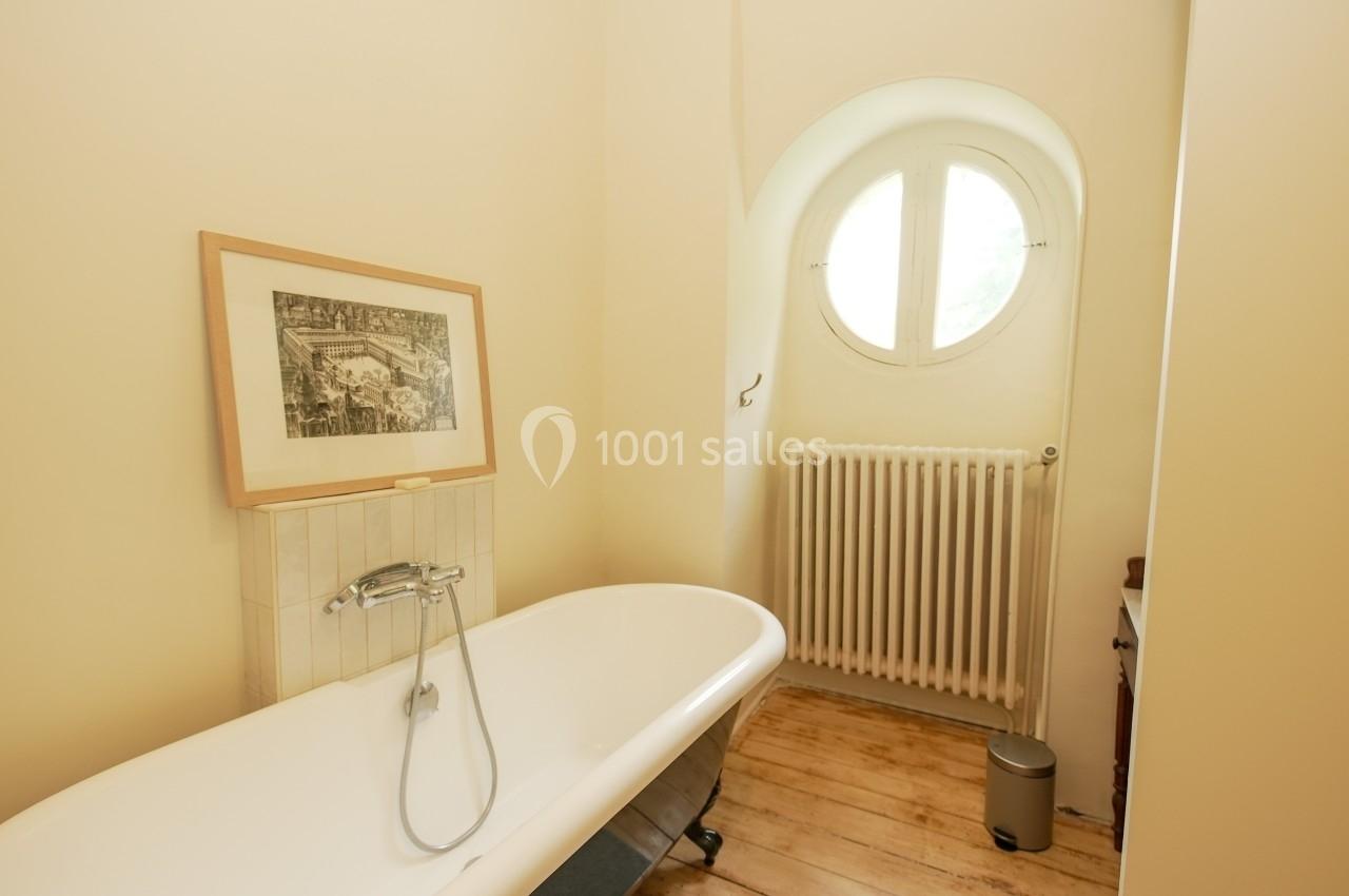 Salle de bain lumineuse avec baignoire sur pieds, radiateur blanc, parquet en bois et fenêtre ronde.