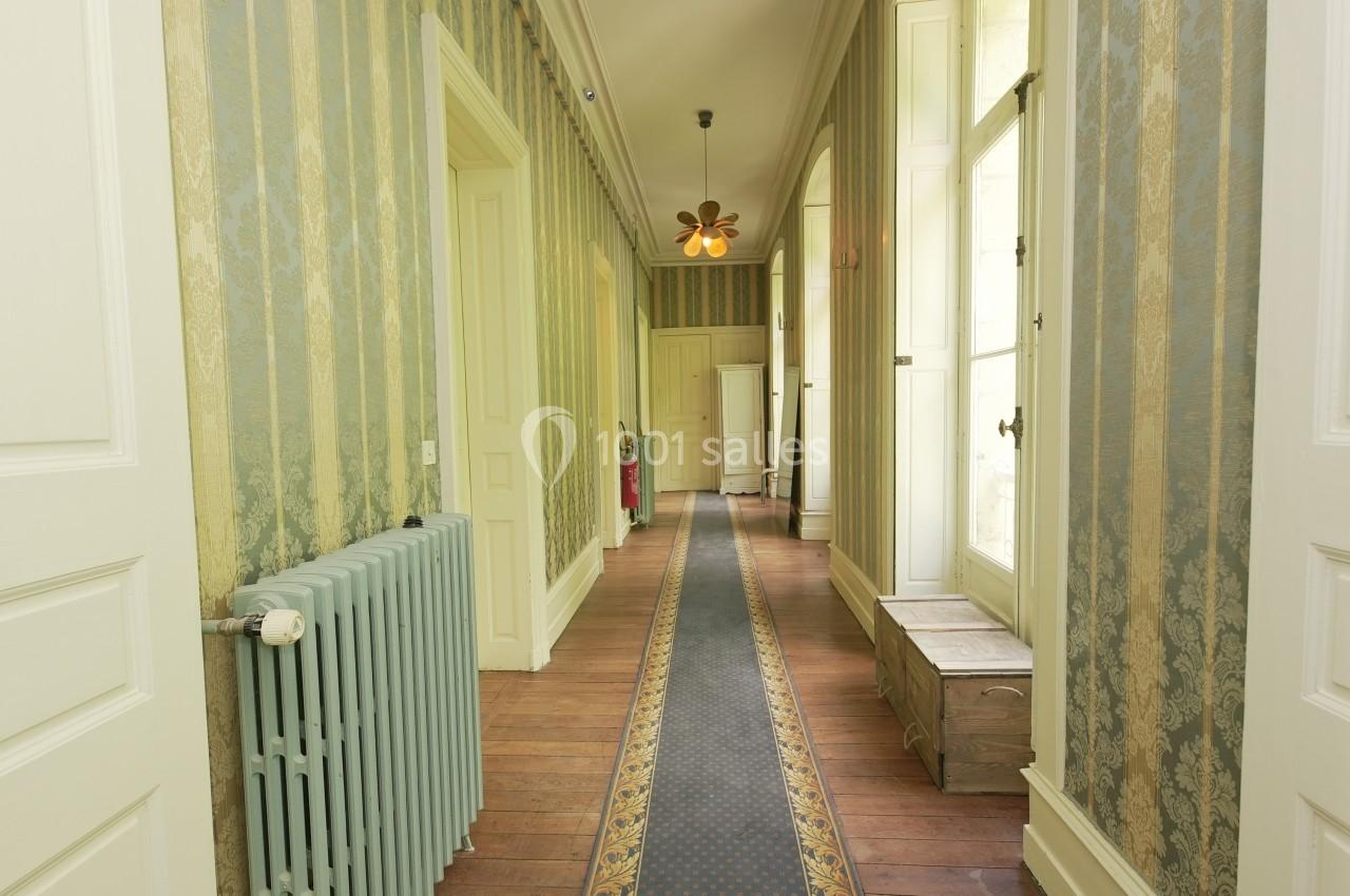 Couloir lumineux avec parquet en bois, tapis central, murs ornés de papier peint et fenêtres laissant entrer la lumière.