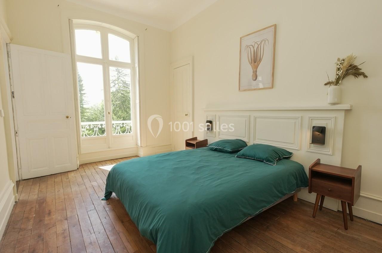 Chambre lumineuse avec un lit double à draps verts, parquet en bois et grande fenêtre donnant sur un espace arboré.