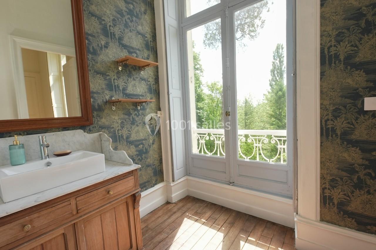 Salle de bain lumineuse avec lavabo en marbre, meuble en bois, papier peint à motifs et vue sur un jardin par une grande…