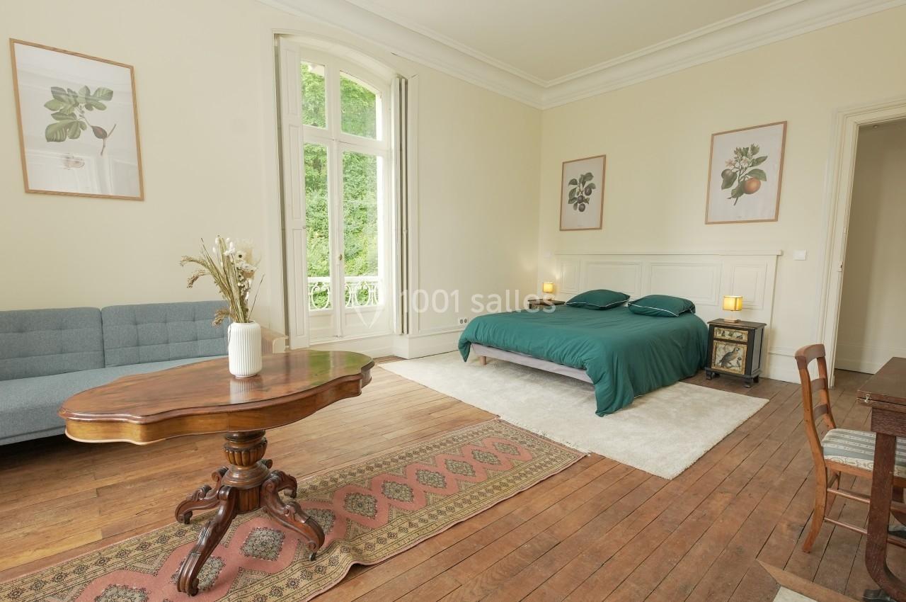 Chambre lumineuse avec lit double, canapé, table en bois, tapis et grandes fenêtres donnant sur un espace vert.