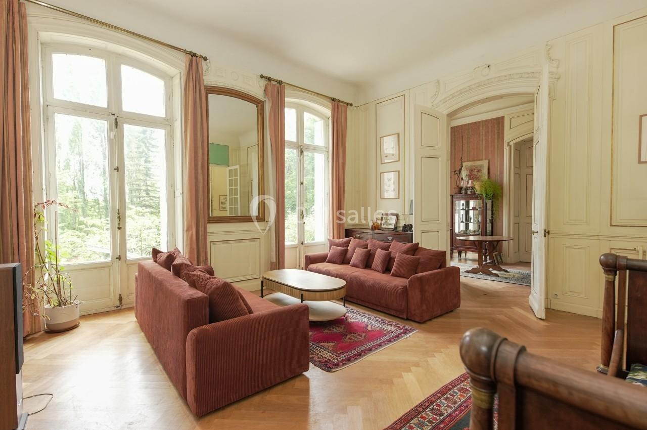 Salon lumineux avec canapés en velours rouge, table basse en bois, grandes fenêtres et parquet en chevrons.