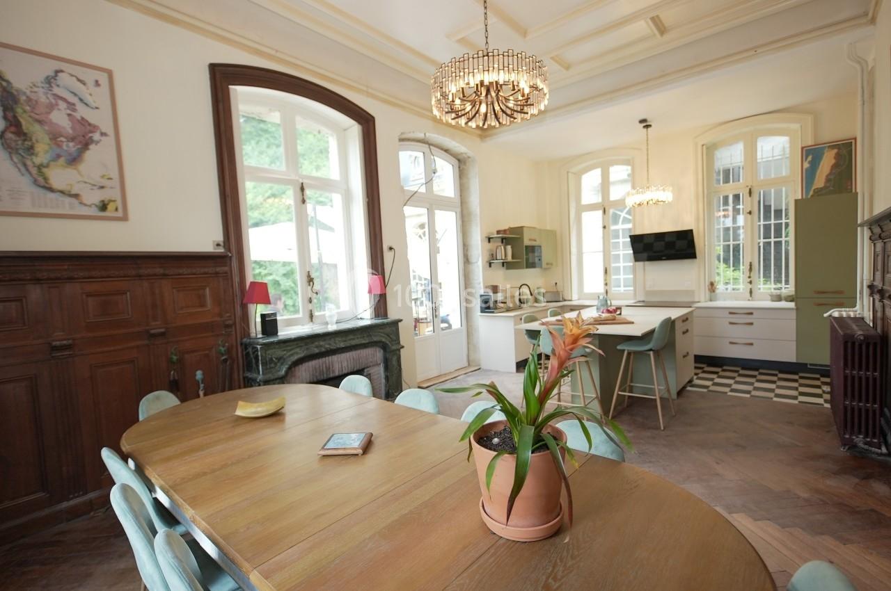 Salle à manger lumineuse avec table en bois, chaises vertes, cuisine ouverte et grandes fenêtres donnant sur un jardin.